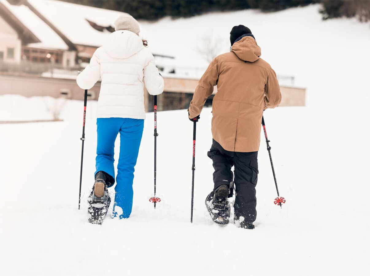 Two people snowshoeing on a snowy landscape, wearing winter gear and using poles, with a building in the background.