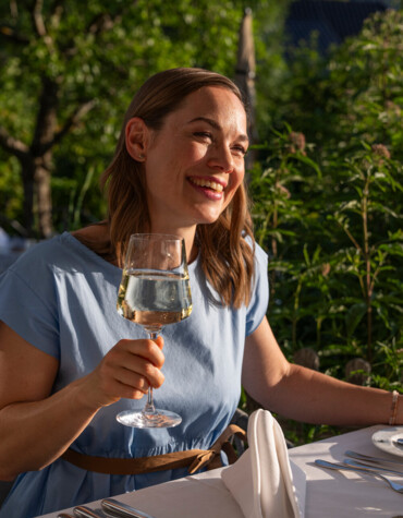 A woman in a blue dress enjoys a glass of wine at an outdoor table surrounded by lush greenery.