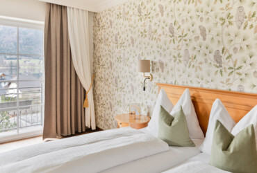 A cozy hotel room with floral and owl wallpaper, a double bed with green pillows, and a scenic view of the lake and mountains through a large window.