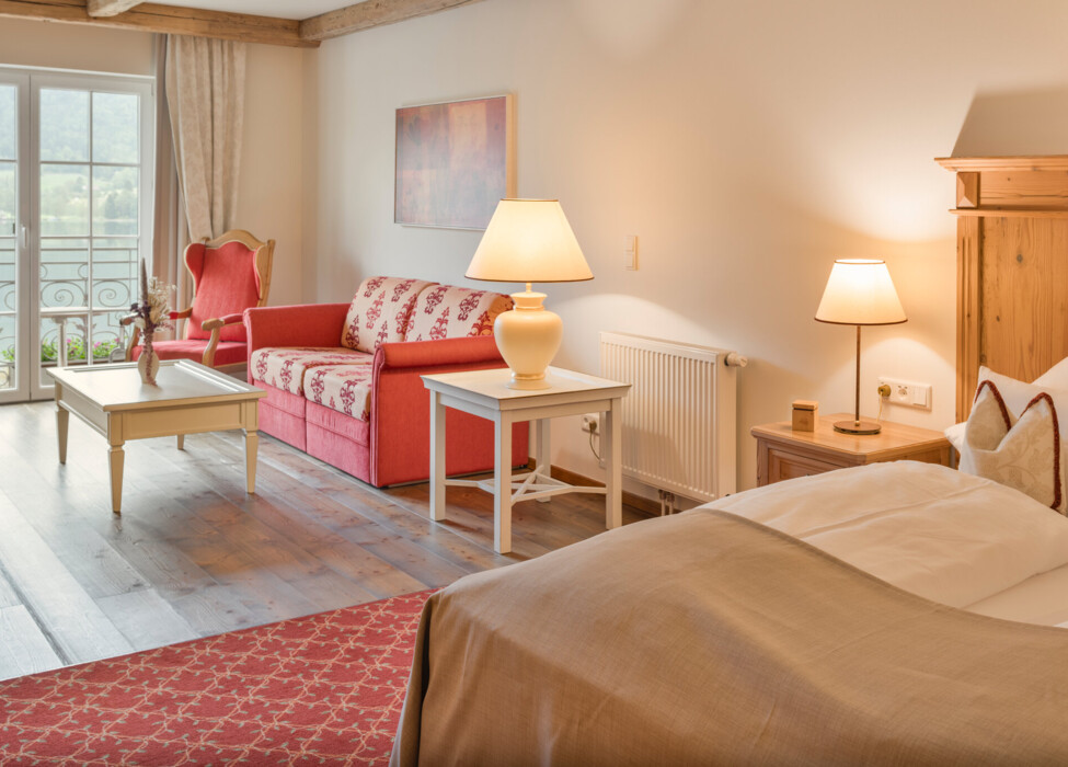 Cozy hotel room with a red sofa, armchair, and warm lighting, featuring a view of the outdoors through French doors.