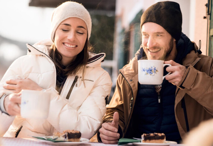 A couple enjoying hot drinks and cake outdoors, dressed warmly in winter clothing.