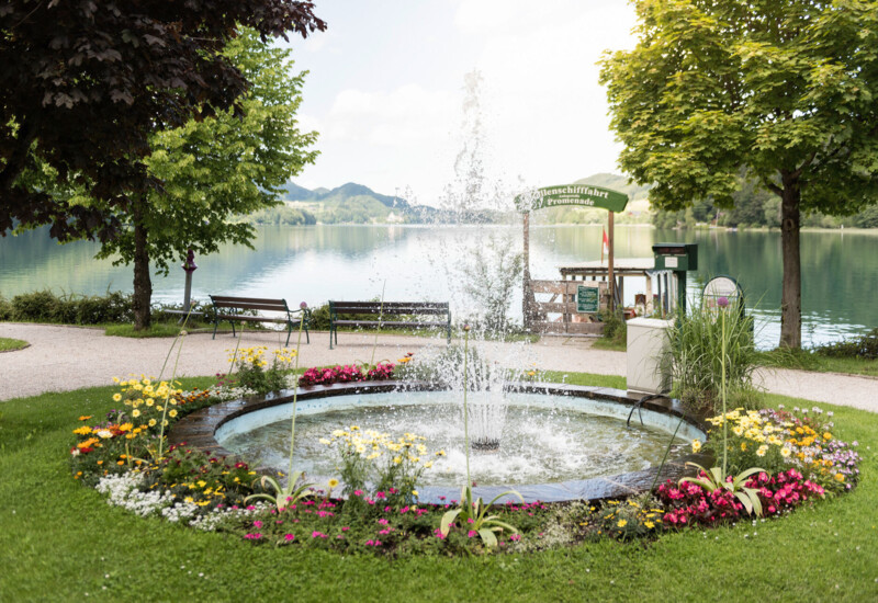 A scenic lakeside view with a lush garden and fountain, surrounded by colorful flowers and trees. Benches offer a relaxing spot by the water.