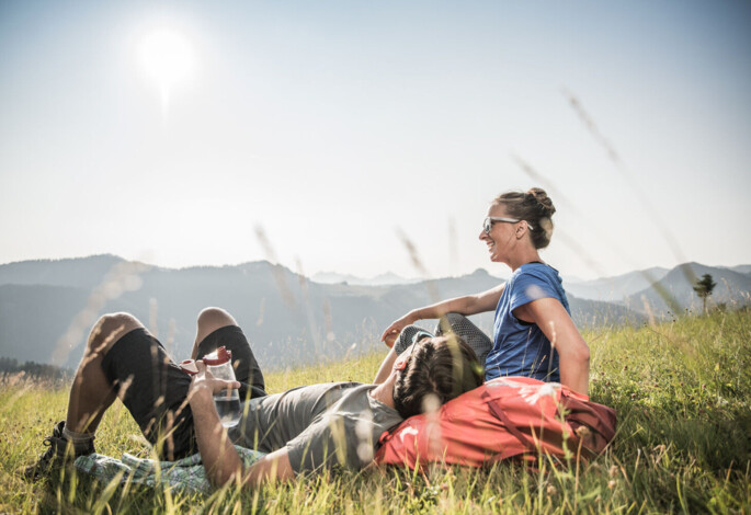 A couple relaxes on a grassy hillside, enjoying a sunny day with scenic mountain views in the background.