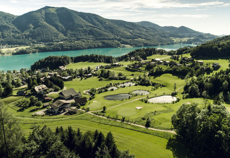 Scenic aerial view of a lush golf course surrounded by mountains and a lake.