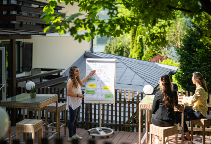 Outdoor meeting with a woman presenting a flip chart to two people, surrounded by lush greenery and a scenic backdrop.