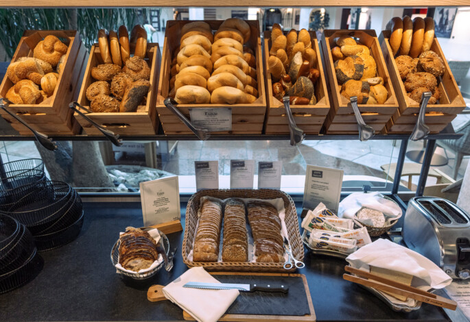 A variety of fresh bread and rolls displayed in wooden trays at a breakfast buffet, with a toaster and condiments nearby.