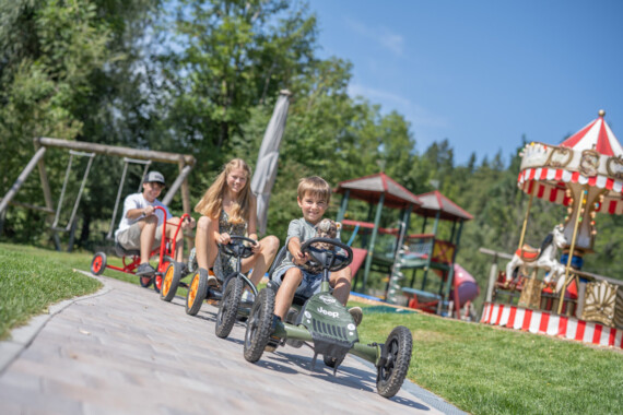 Children enjoying go-karting at Waldhof Fuschlsee Resort with a carousel and playground in the background.