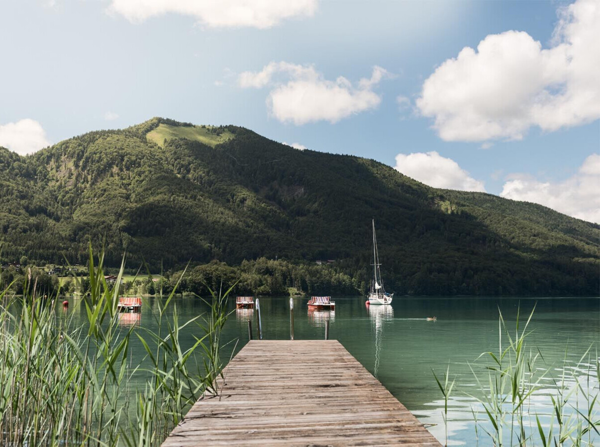 A wooden pier extends into a tranquil lake surrounded by lush mountains under a partly cloudy sky. Sailboats are anchored nearby, creating a scenic view.