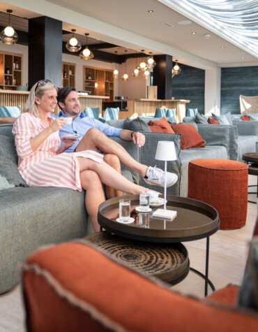 A couple relaxes on a stylish couch in a modern, well-lit lounge with elegant decor and comfortable seating.