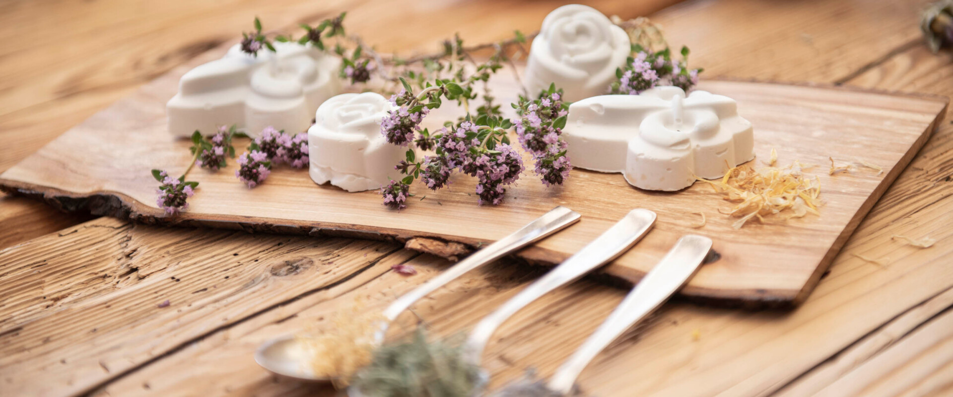 Handcrafted soaps with herbs on a wooden board, accompanied by spoons with dried flowers.