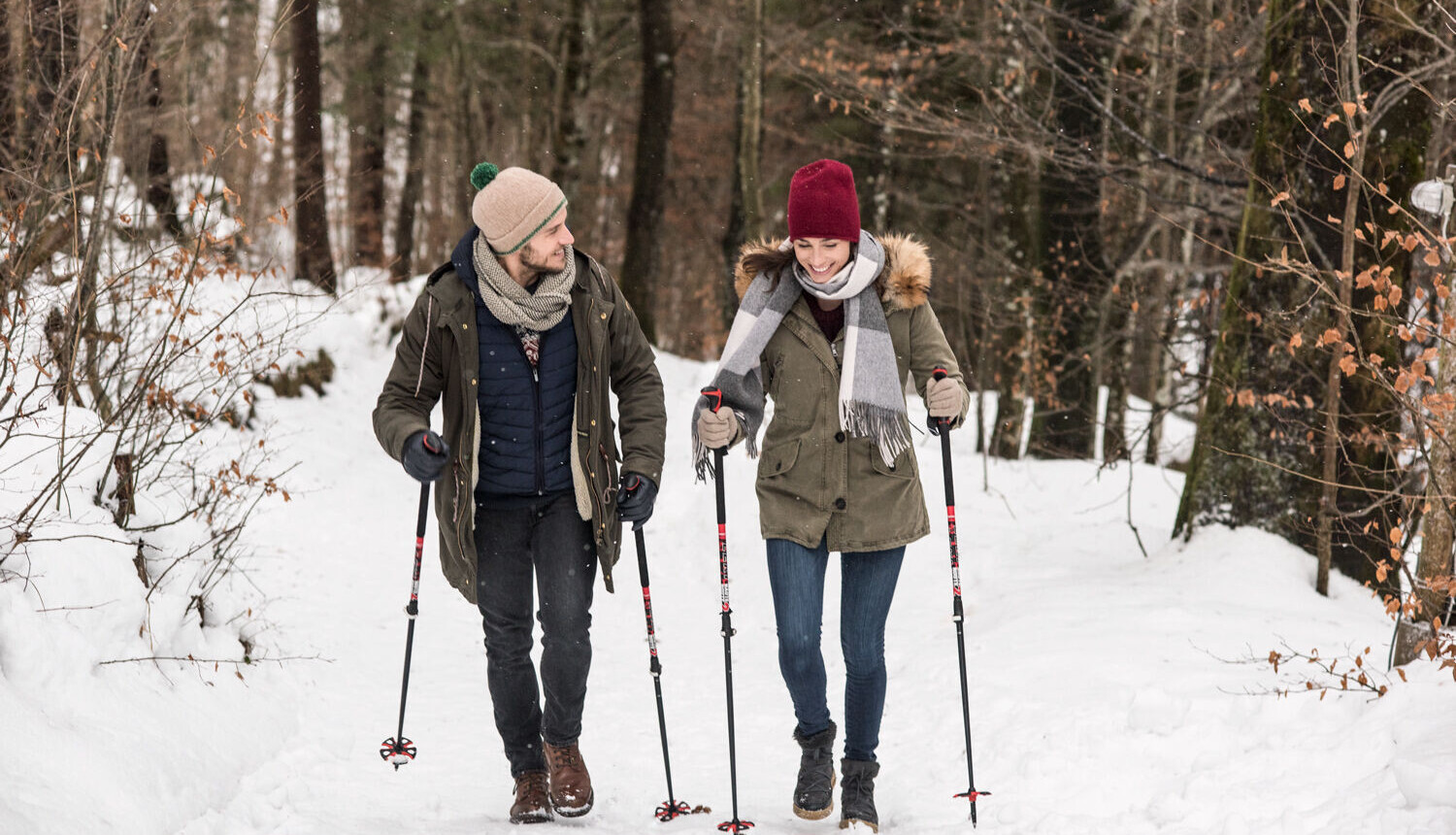 A couple enjoys a winter hike through a snowy forest, using trekking poles for support.