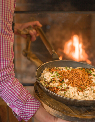 Ein Koch in kariertem Hemd hält eine Pfanne mit frisch zubereiteten Käsespätzle vor einem offenen Kamin.