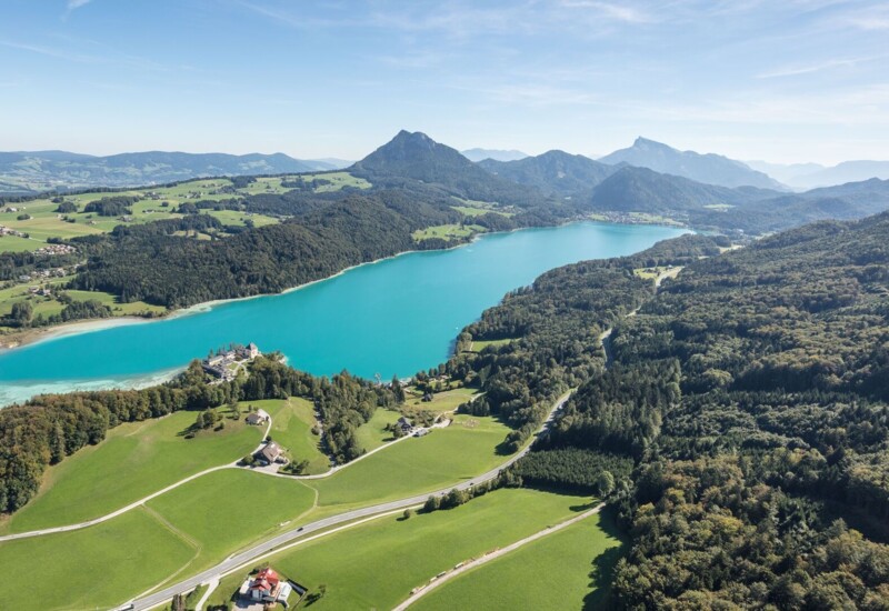 Aerial view of a scenic lake surrounded by lush greenery and mountains under a clear blue sky.