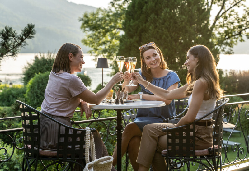 Three women toasting with champagne at an outdoor table, surrounded by lush greenery and a scenic lake view.