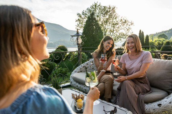 Drei Frauen genießen Drinks auf einer Terrasse mit Blick auf die Natur bei Sonnenuntergang.