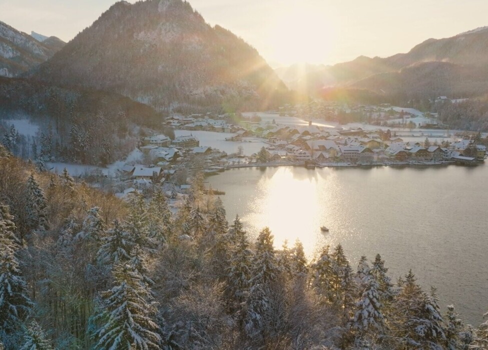 Snow-covered landscape with sunlit mountains and a shimmering lake, surrounded by frosted trees and a quaint village in the distance.