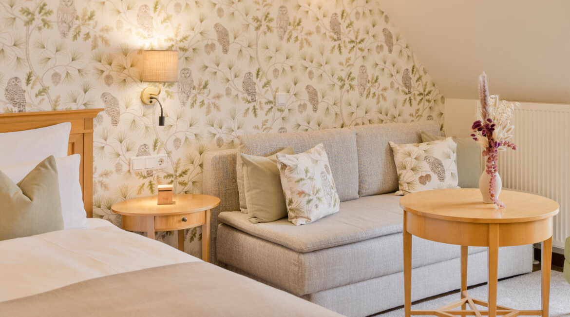 Cozy hotel room with floral wallpaper, a bed, a sofa with patterned cushions, and a round wooden table with a vase.