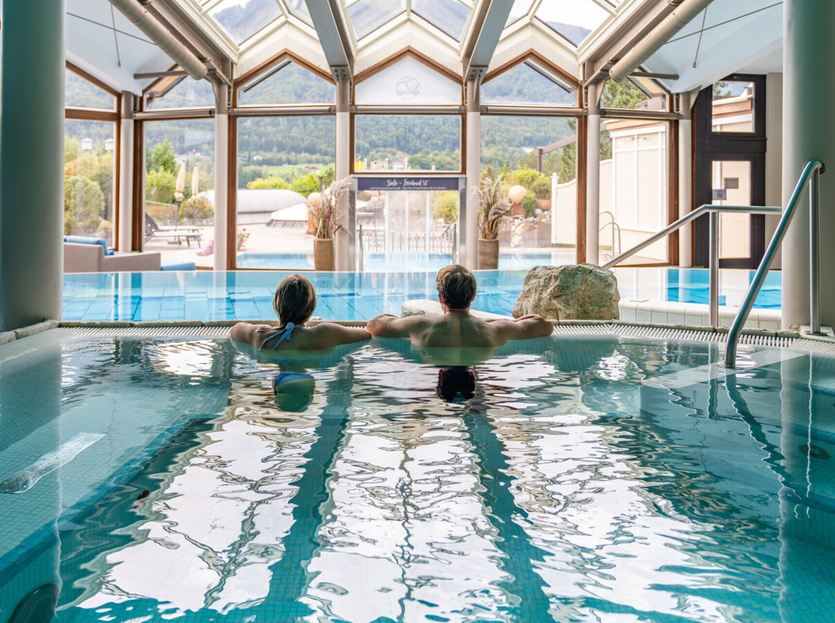 Couple relaxing in an indoor pool with scenic mountain views through large windows.