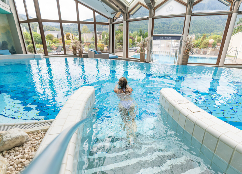 A person swims in an indoor pool with large windows at Waldhof Fuschlsee Resort, offering views of the surrounding landscape.