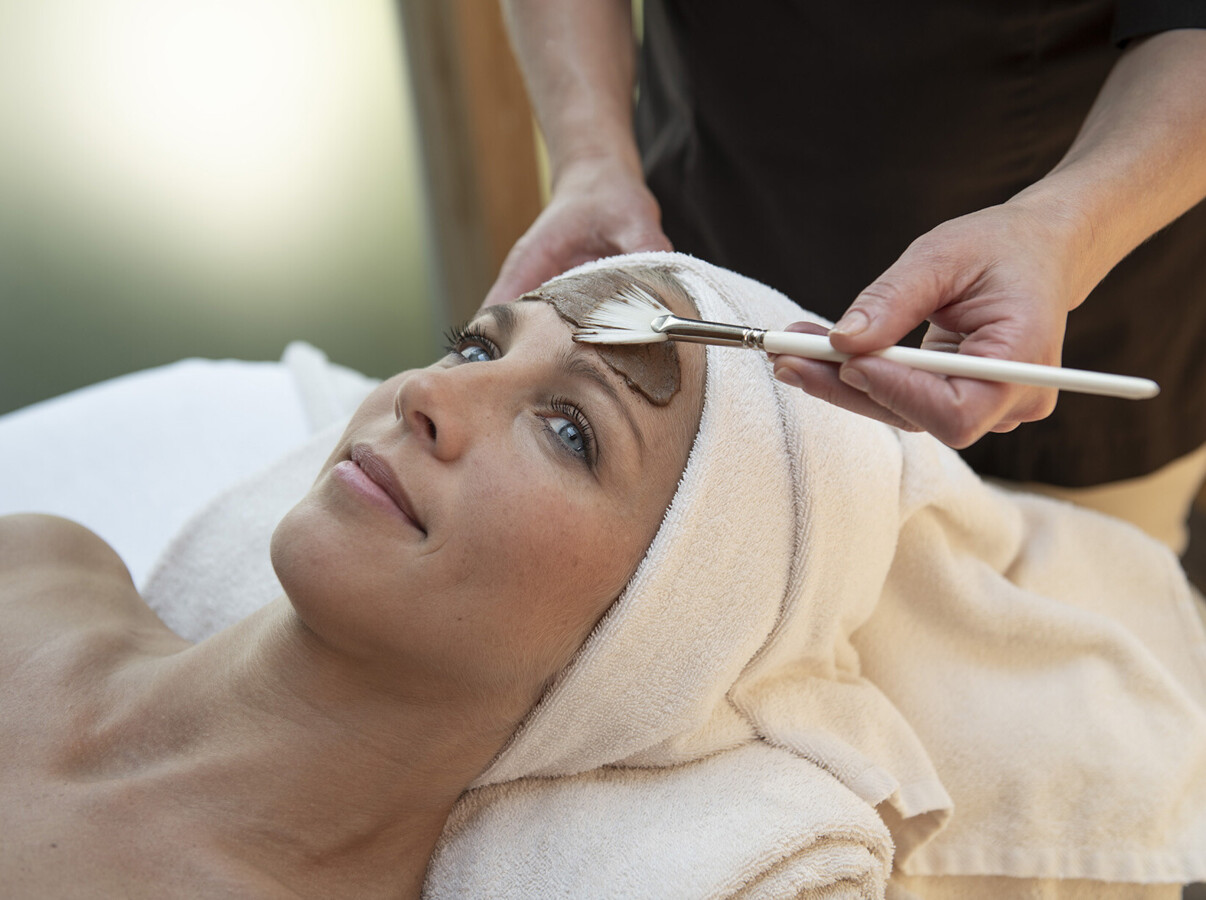 A woman receives a relaxing facial treatment with a brush applicator at a spa, wrapped in towels.