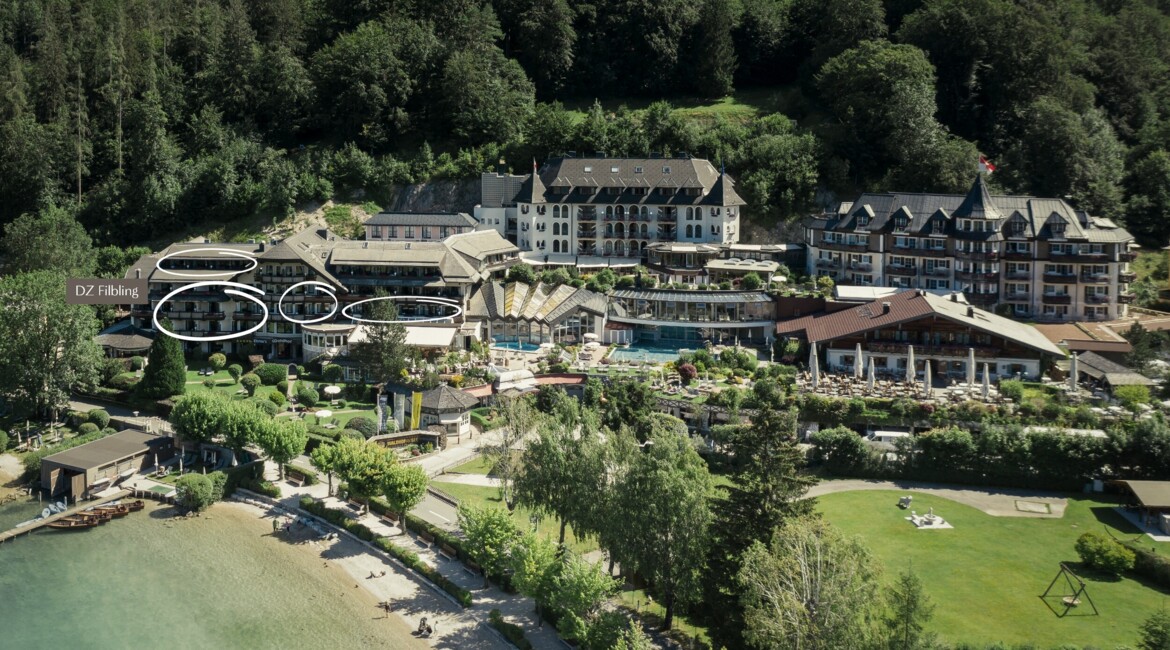 Luftaufnahme des Walhof Fuschlsee Resorts, umgeben von Wald und Gartenanlagen. Im Vordergrund sind der See und ein kleiner Strand sichtbar.