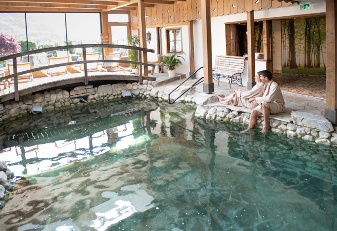 Couple in robes sitting by an indoor pool at Waldhof Fuschlsee Resort, with lounge chairs and stone decor.