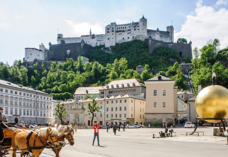 Festung Hohensalzburg überragt den Residenzplatz in Salzburg mit Pferdekutsche und Kunstinstallation im Vordergrund an einem sonnigen Tag.