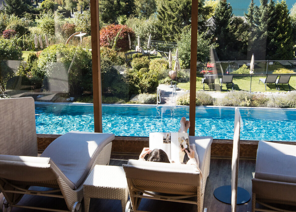 Eine Person entspannt auf einer Liege am Pool mit Blick auf Berge und einen See durch eine Glasfront.