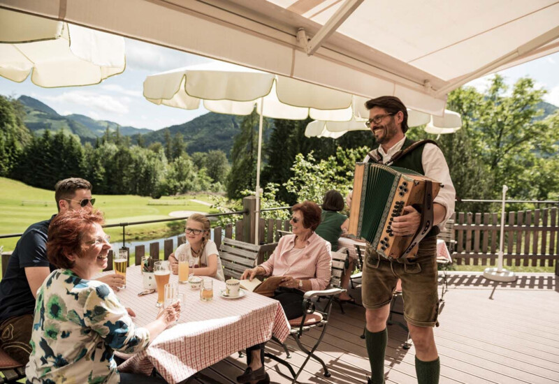 A family enjoys drinks on a terrace with scenic mountain views, as a musician plays the accordion.