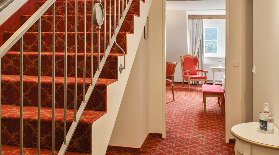 Red-carpeted staircase and hallway leading to a brightly lit room with chairs and a window.