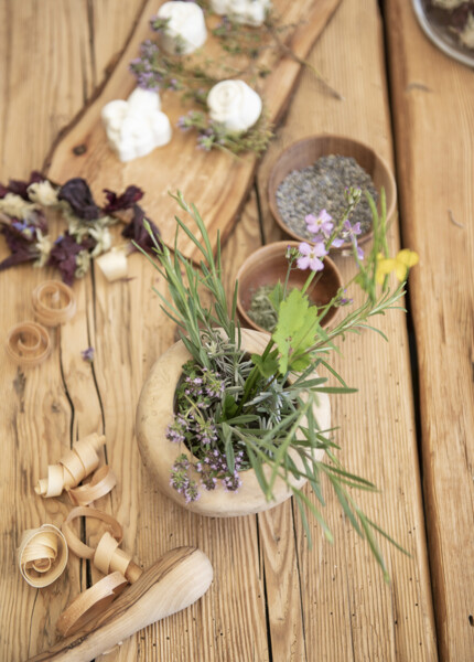 Herbs and flowers in a wooden bowl on a rustic table, surrounded by dried petals and wooden accents.