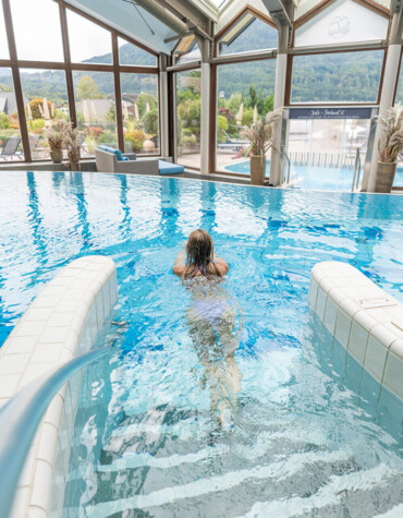 Eine Person schwimmt im Innenpool mit Blick auf die Berge durch große Fenster.