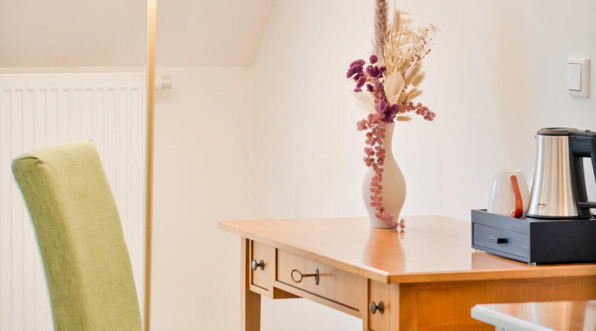 A cozy room corner with a wooden desk, green chair, floor lamp, and a vase of dried flowers, creating a warm, inviting atmosphere.