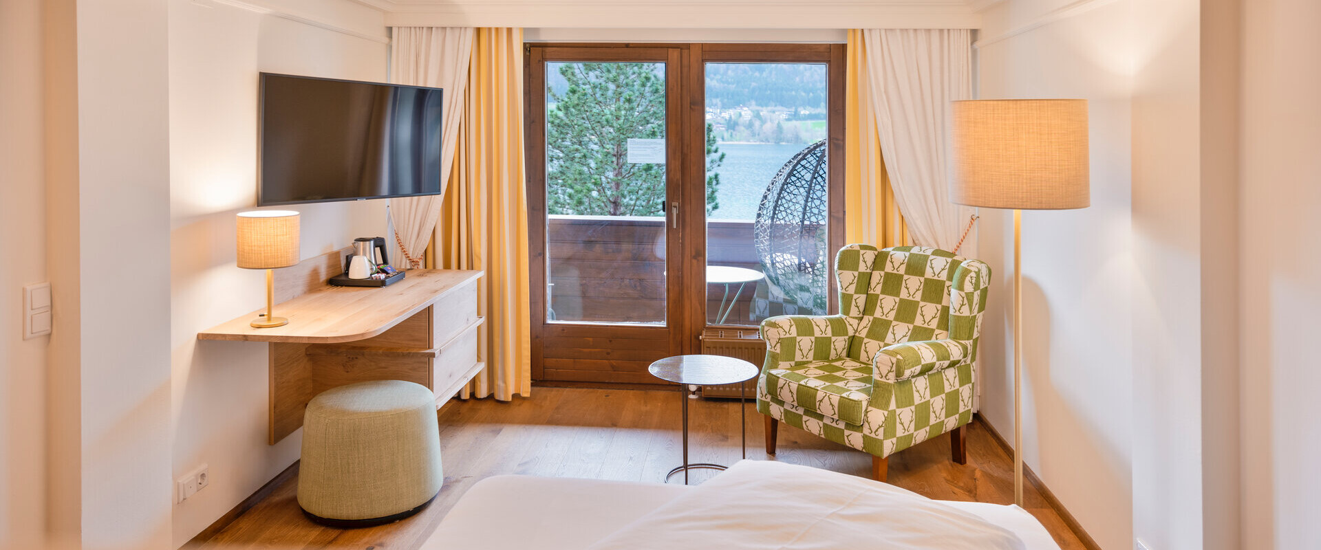 Elegant hotel room with a cozy seating area, large TV, and balcony overlooking a serene lake, offering a tranquil retreat.