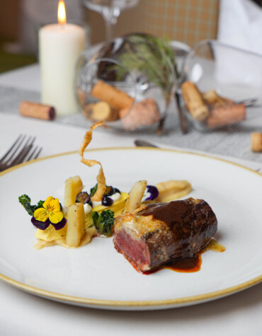 Elegant dish with meat and vegetables, garnished with flowers, on a fine dining table setting with candlelight and wine corks.