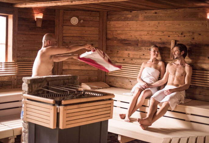 A sauna with a man enhancing heat while a couple relaxes on benches.