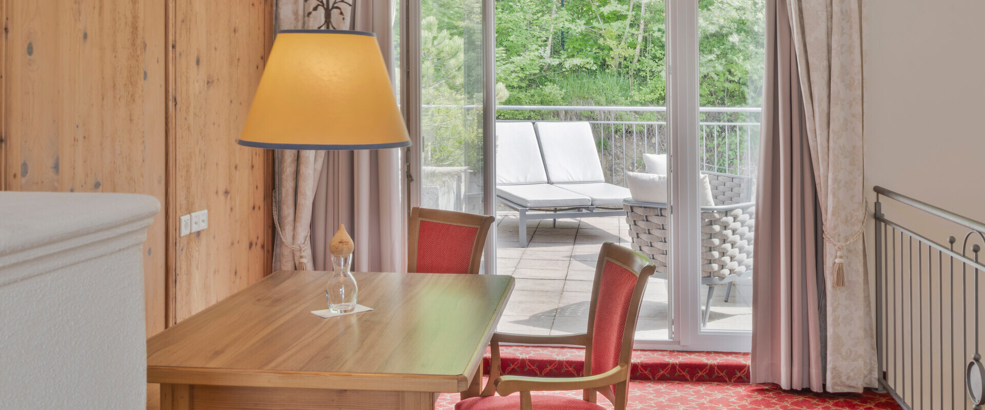 Cozy room with wooden table and red chairs, overlooking a lush balcony with sun loungers through glass doors.