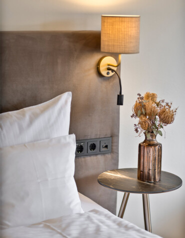 Bedside area with plush headboard, modern wall lamp, and a small table holding a vase with dried flowers at Waldhof Fuschlsee Resort.