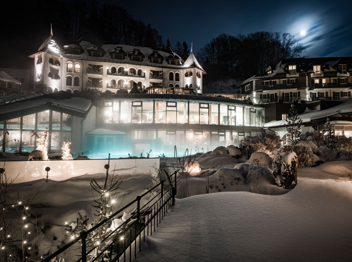 Snow-covered hotel at night, warmly lit with decorative lights and a glowing pool, under a full moon.