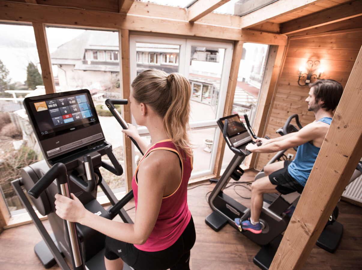 People exercising on machines in a wooden fitness room with scenic views outside.