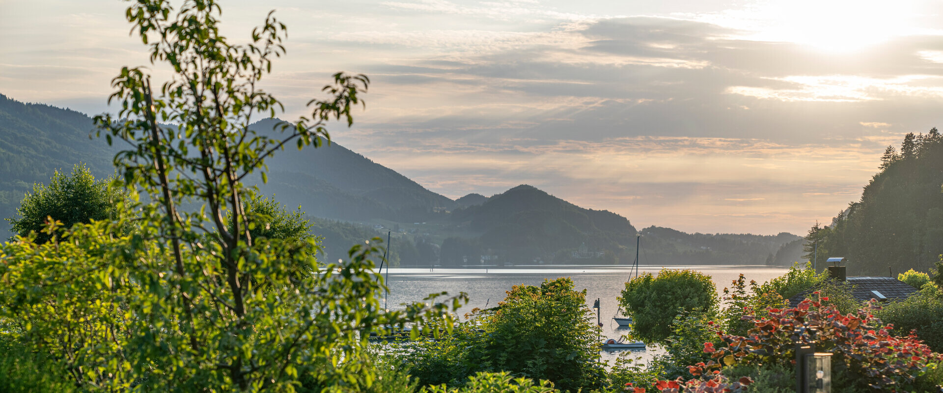 Scenic view of a lush garden overlooking a tranquil lake at sunset, with mountains in the background.