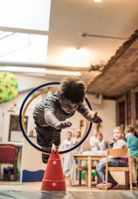 A child jumps through a hoop during an indoor activity at a play center, with other children watching in the background.