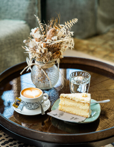 Cup of coffee, slice of cake, and glass of water on a wooden table with dried floral arrangement in a cozy setting.