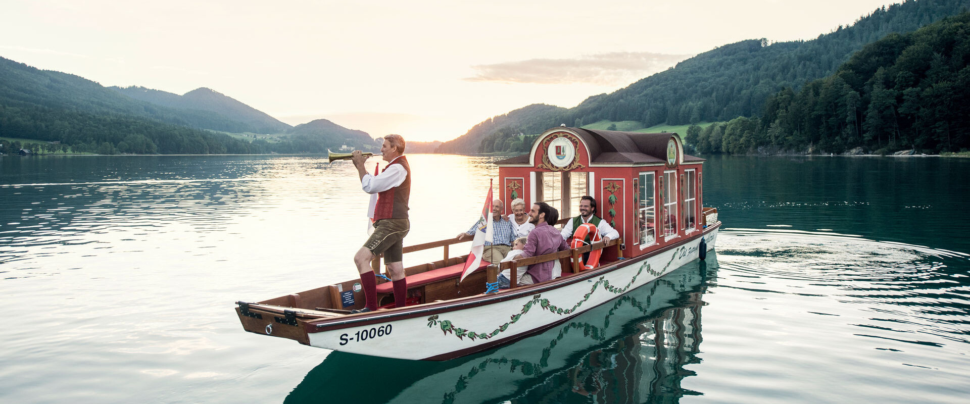 A boat with people in traditional attire sails on a serene lake surrounded by lush, scenic mountains at sunset.