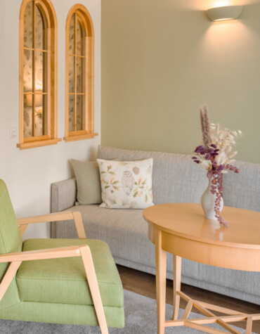 A cozy living area with a green armchair, gray sofa, wooden table, and decorative vase. Two arched windows add charm to the space.