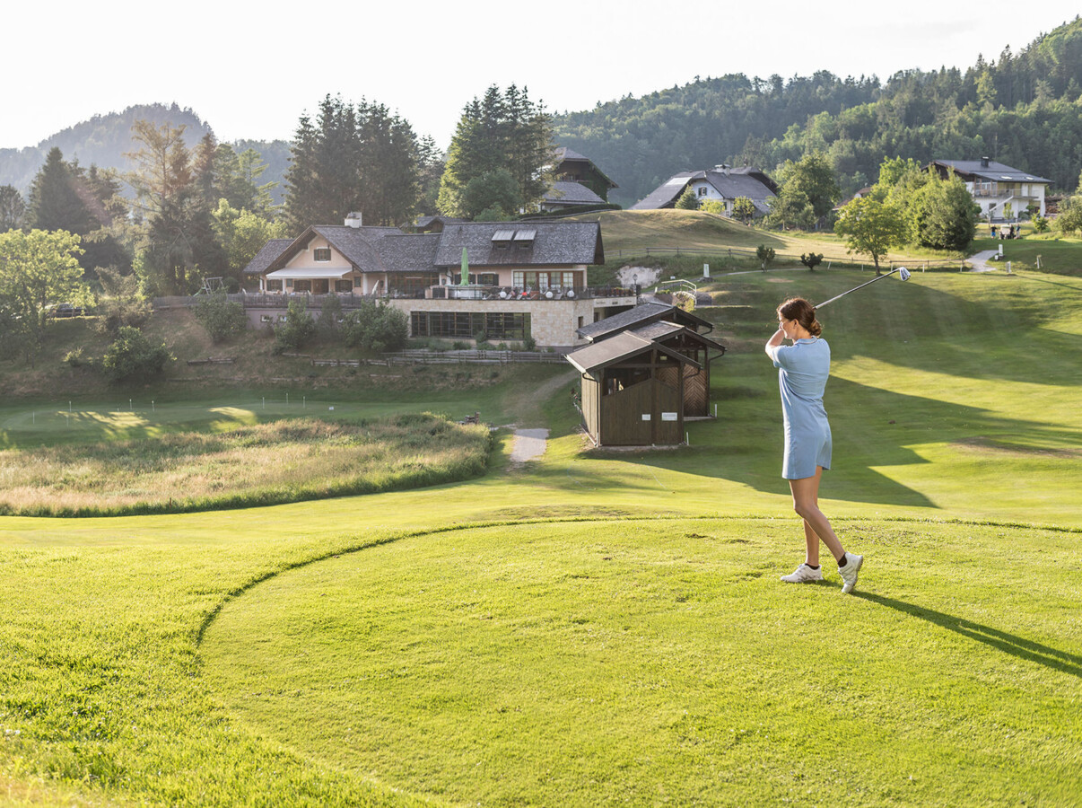 A woman playing golf on a lush, scenic course with picturesque buildings and hills in the background.