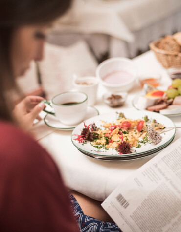 A person enjoys breakfast with a newspaper, featuring a plate of scrambled eggs and a variety of foods on a table.