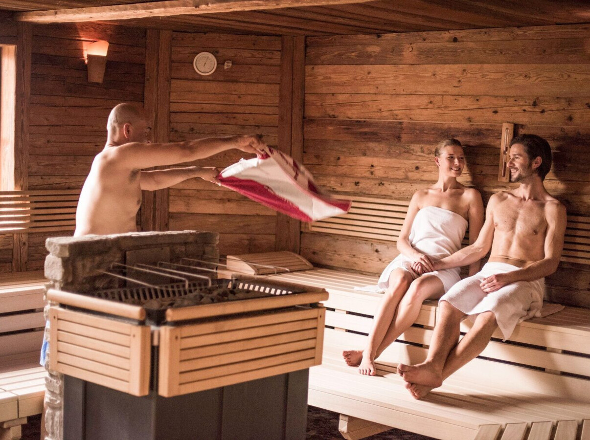 A sauna with a man enhancing heat while a couple relaxes on benches.