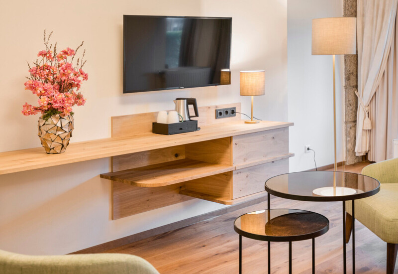 A cozy hotel room with a wall-mounted TV, decorative vase with flowers, and modern furnishings, including a lamp and round tables.