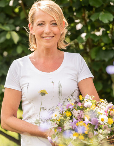 A person smiling while holding a basket of colorful flowers, with a lush green background.